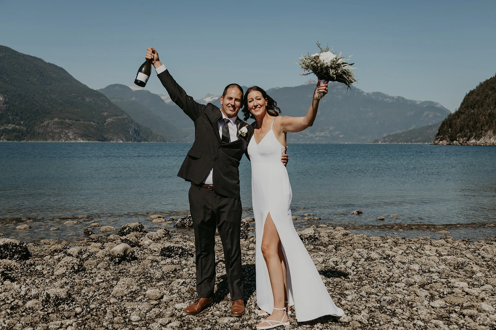 Bride and groom celebrate after their ceremony by holding up a bottle of champagne and a bouquet
