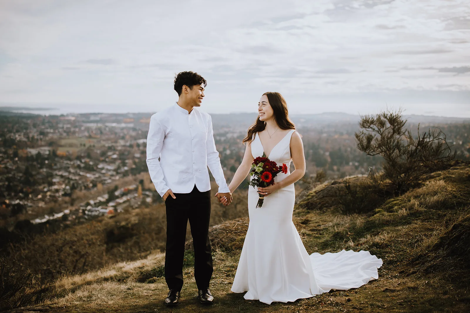 Bride and groom hold hands and smile at each other with a city view behind them