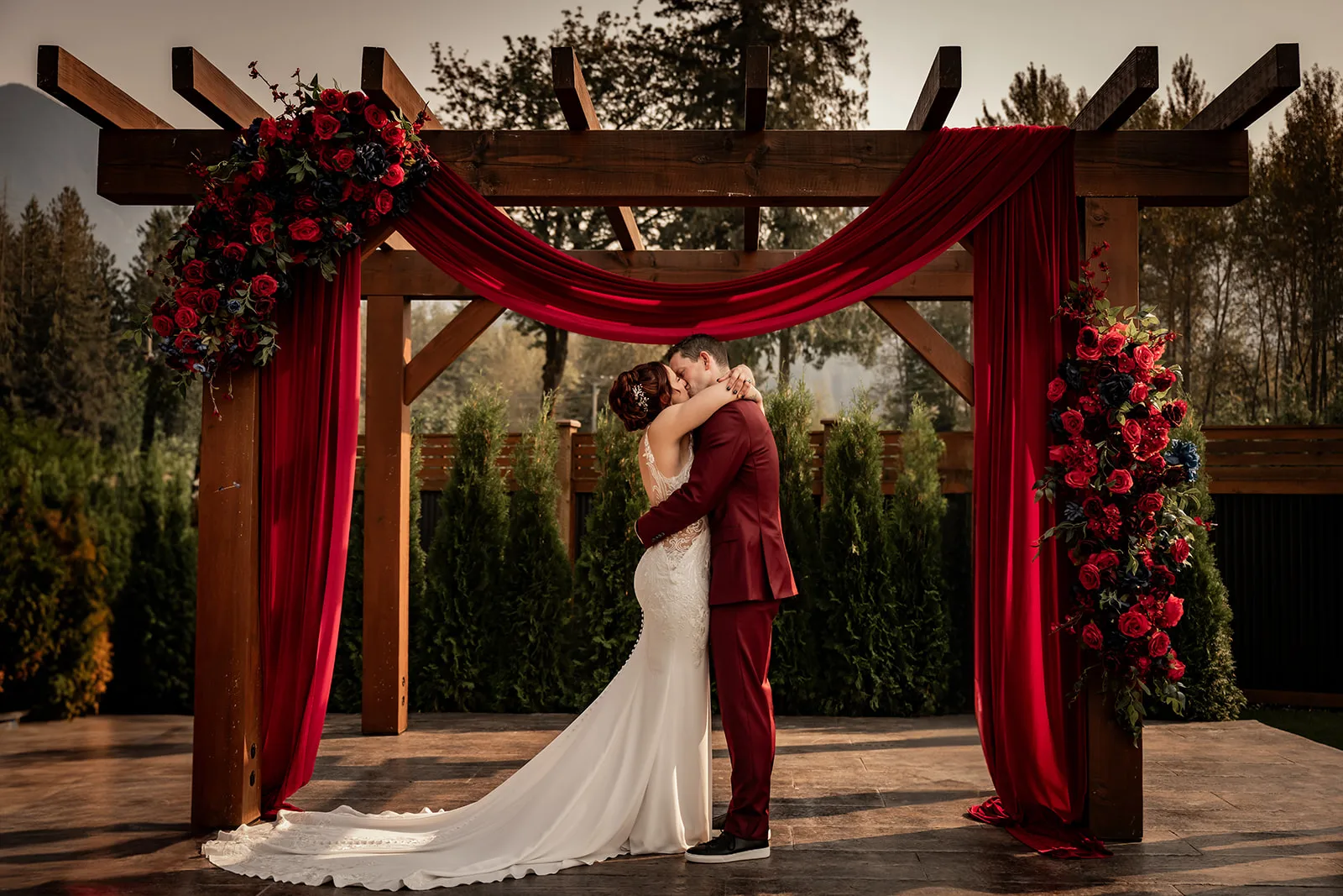 Jennifer and Sean share their first kiss at the end of their wedding ceremony in front of a wooden pergola draped in red curtains, Candace Fast Photography