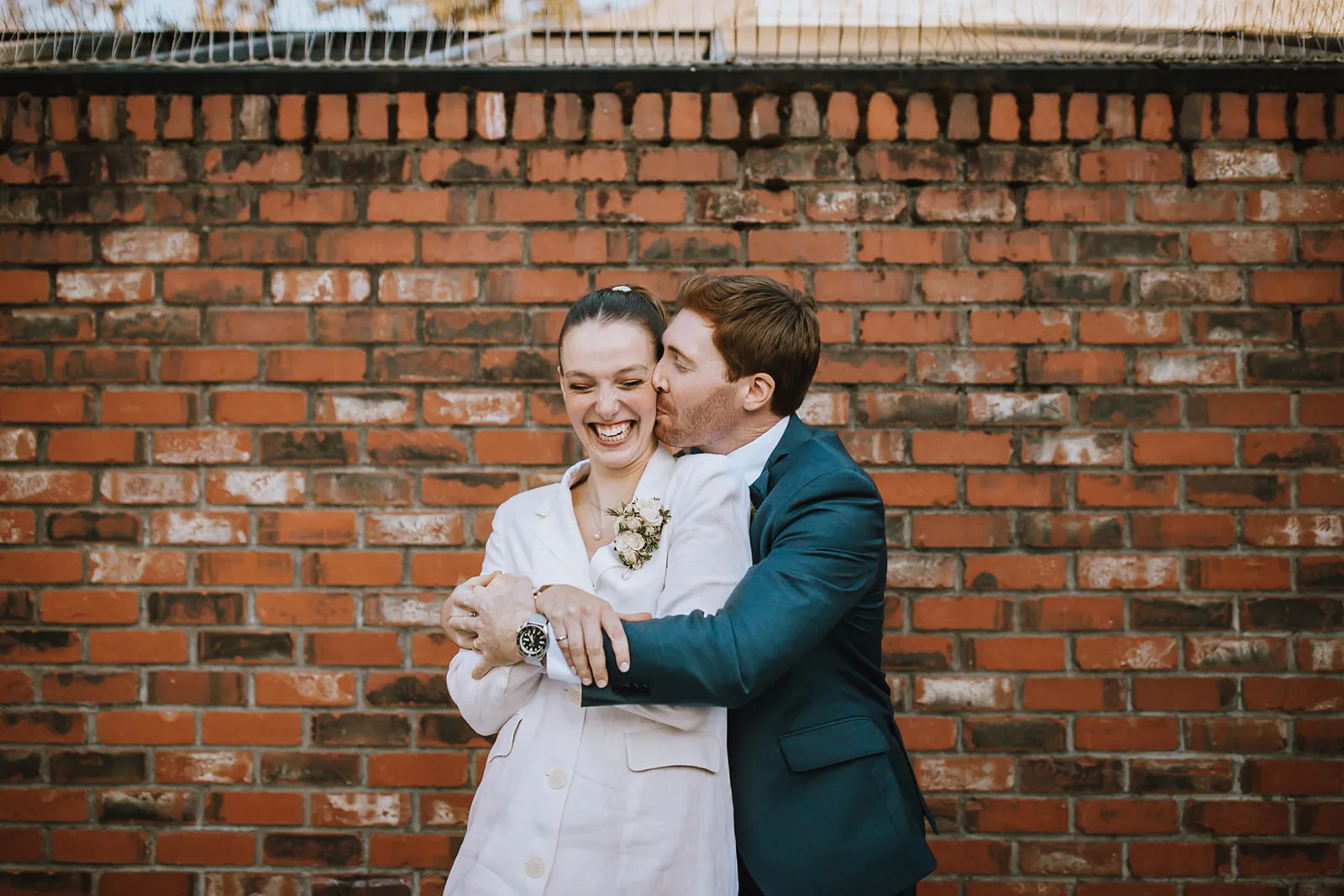 Newlywed couple in front of brick wall where groom plants a kiss on the bride's cheek; Jades Photography